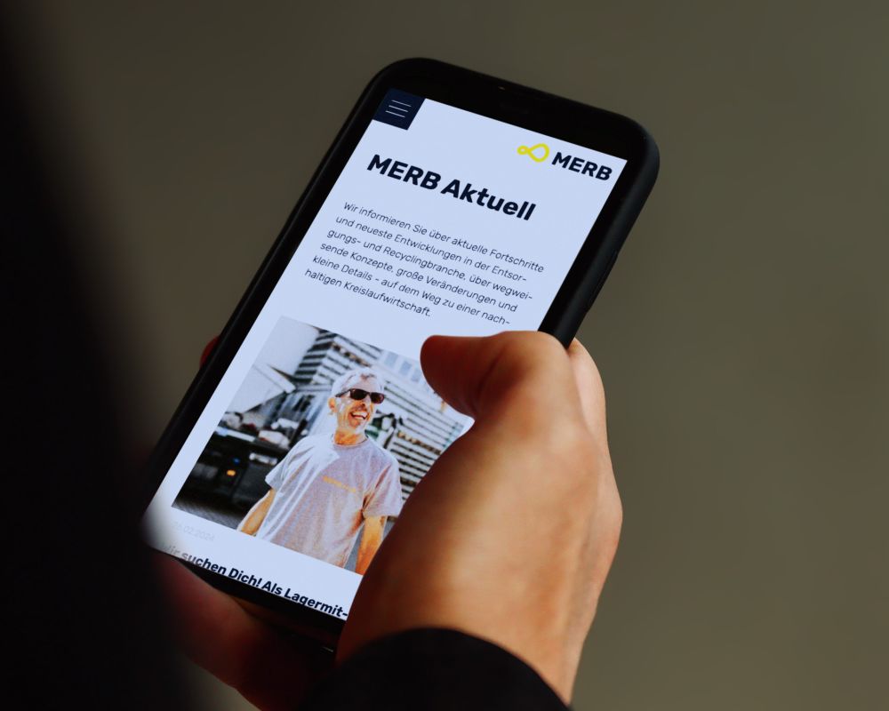 MERB-News auf dem Smartphone Hand hält Smartphone mit geöffneter MERB-Webseite, die aktuelle Informationen und ein Bild eines Mitarbeiters zeigt.