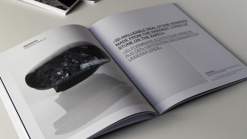 LIXOS Broschüre mit 3D-Steinobjekt Aufgeschlagene LIXOS-Broschüre zeigt 3D-formbares Natursteinfurnier mit Text und dunkel glänzendem Steinobjekt auf weißem Tisch.